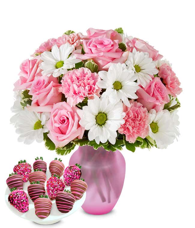 Deluxe bouquet of pink roses, pink carnations, and white daisy poms in a clear glass vase, paired with chocolate-covered strawberries decorated with pink drizzle and pink sprinkles.