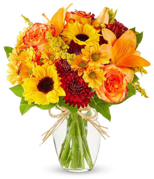A vibrant fall bouquet in clear glass vase, sunflowers and orange lilies and roses are surrounded by yellow and maroon filler flowers in a clear glass vase.