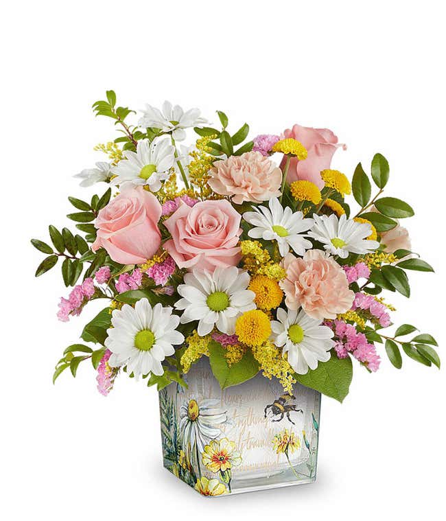 Pink roses, light peach carnations, white daisy spray chrysanthemums, yellow button spray chrysanthemums, pink statice, solidago, and floral greenery arranged in a square glass container featuring a watercolor-style bee and floral design.
