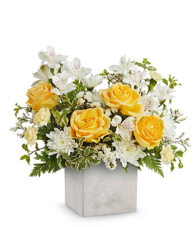 A floral arrangement in a square gray container featuring yellow roses, white alstroemeria, white carnations, white poms, ivy, and seasonal greenery.