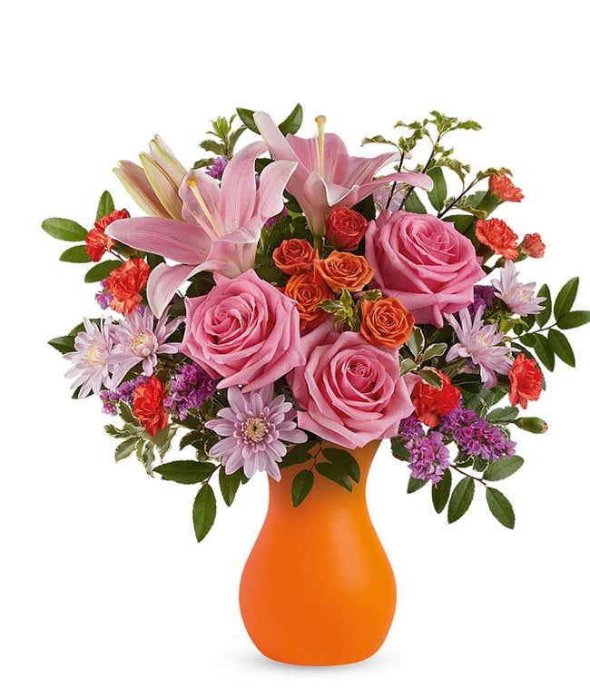 Vibrant floral arrangement featuring pink roses, pink Asiatic lilies, orange spray roses, red miniature carnations, lavender cushion spray chrysanthemums, purple statice, and lush greenery in a bold orange vase.