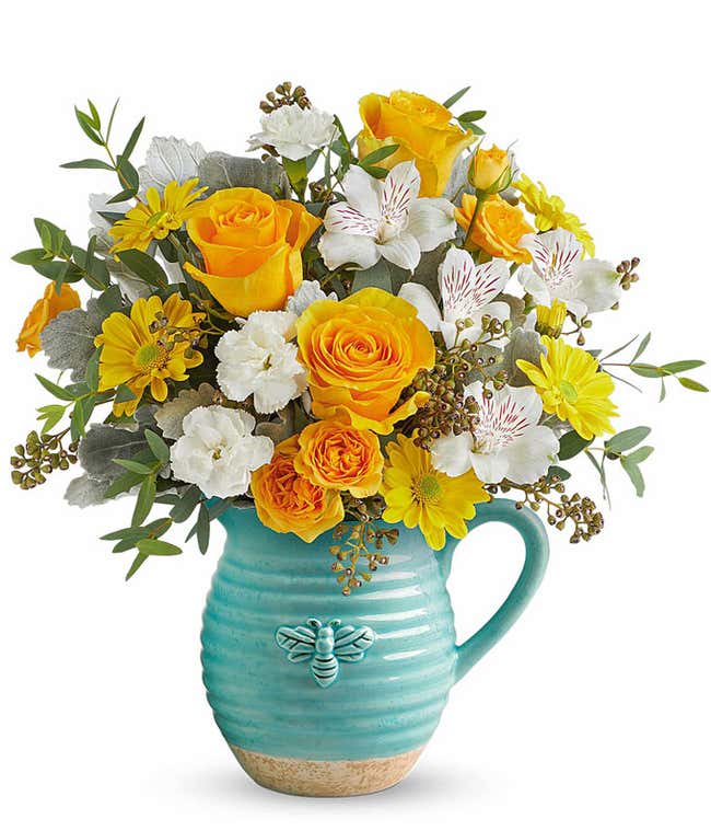 Yellow roses, white alstroemeria, miniature white carnations, yellow daisy poms, and greenery arranged in a teal ceramic pitcher with a bee design.