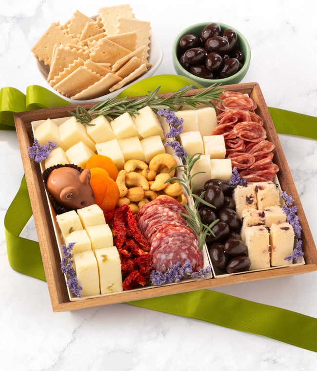 Charcuterie box with cubed cheeses, salami slices, chocolate-covered almonds, dried apricots, cashews, sun-dried tomatoes, shortbread with cranberries, a chocolate mouse, fresh rosemary, and purple flower accents.